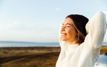 A woman in a white sweater and brown beanie stands with eyes closed, smiling contentedly with arms raised behind her head. She enjoys a sunny day by a beach, with a clear blue sky and the sea in the background.