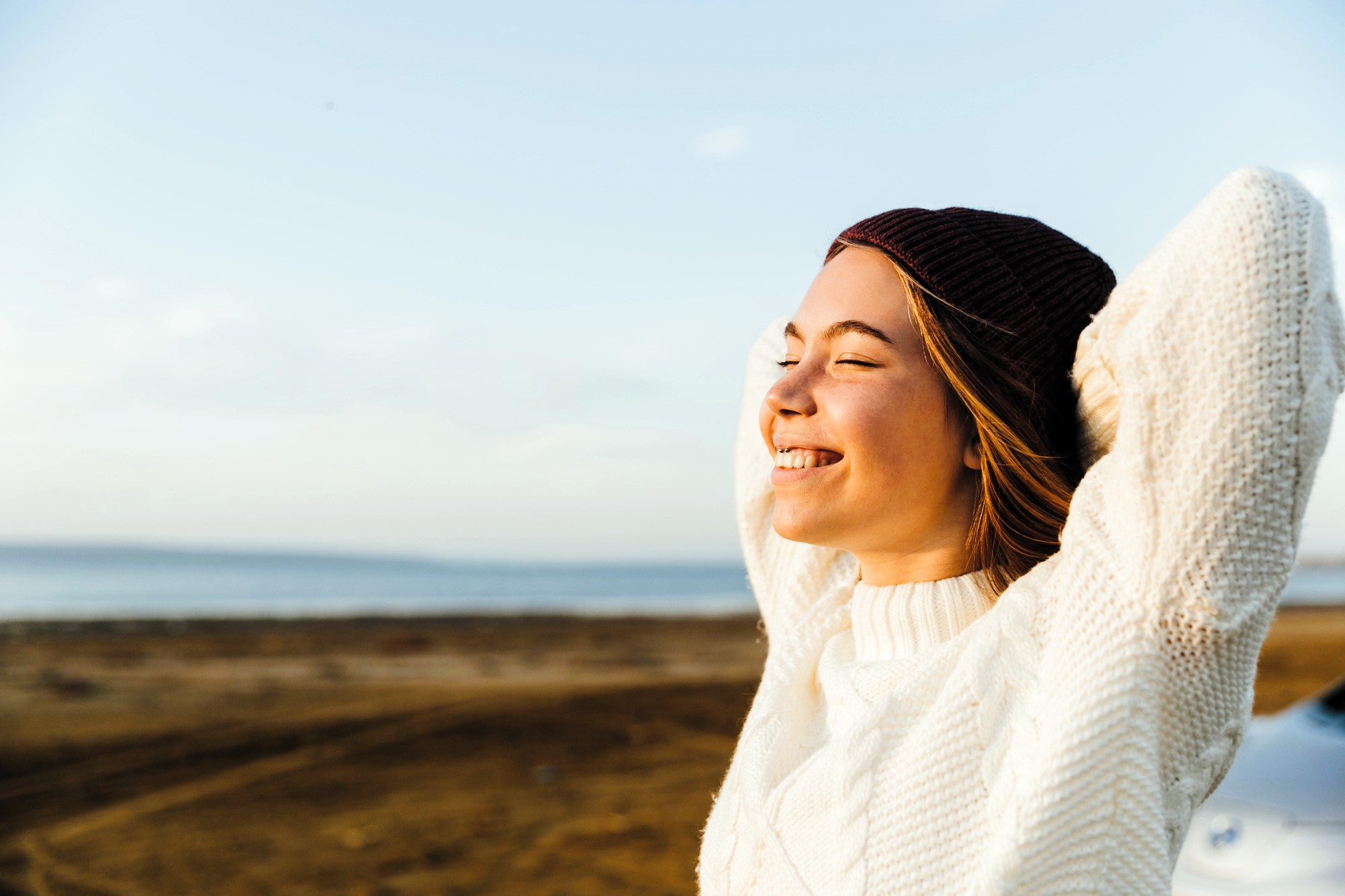 A woman in a white sweater and brown beanie stands with eyes closed, smiling contentedly with arms raised behind her head. She enjoys a sunny day by a beach, with a clear blue sky and the sea in the background.