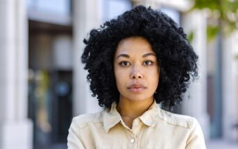 Woman with curly black hair, wearing a beige collared shirt, stands with arms crossed in front of a blurred building background. She looks directly at the camera with a neutral expression.