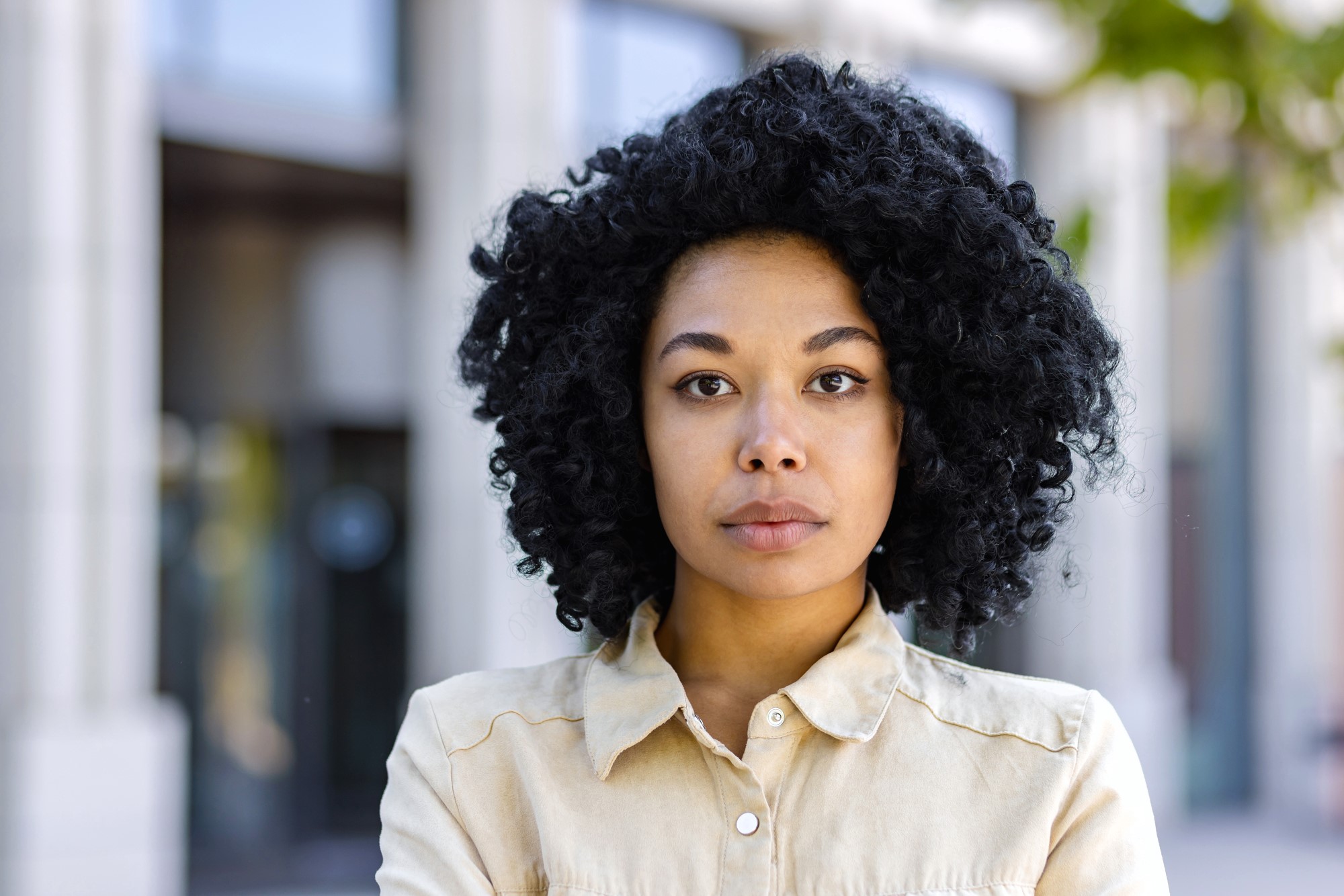 Woman with curly black hair, wearing a beige collared shirt, stands with arms crossed in front of a blurred building background. She looks directly at the camera with a neutral expression.