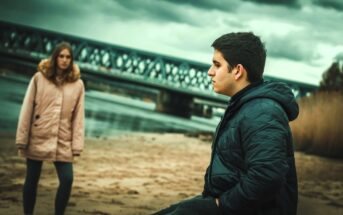 A young man in a dark jacket stands on a sandy beach, looking pensive. In the background, a woman in a light-colored coat stands near a river, with an overcast sky and a metal bridge visible.