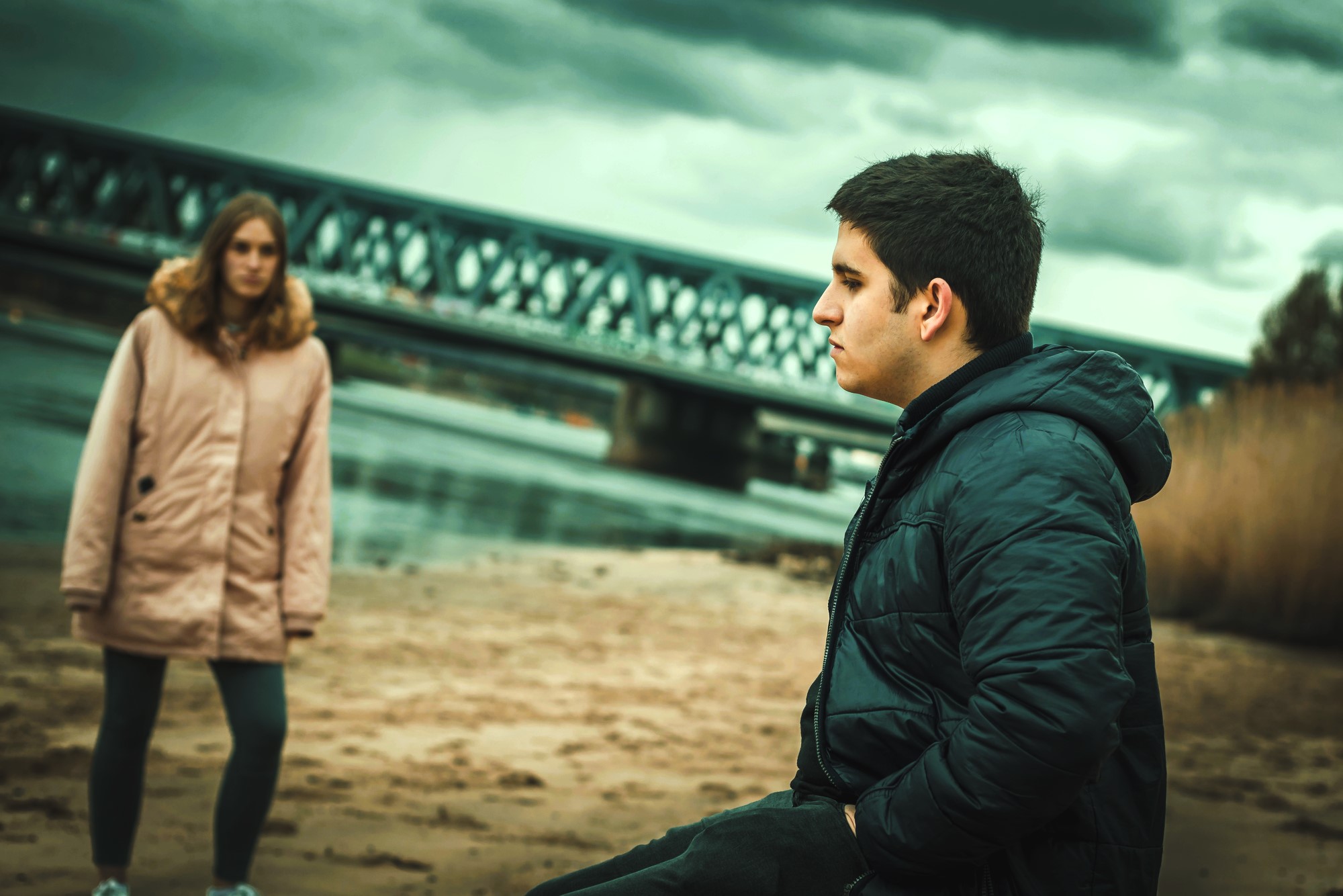 A young man in a dark jacket stands on a sandy beach, looking pensive. In the background, a woman in a light-colored coat stands near a river, with an overcast sky and a metal bridge visible.