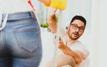 A man sitting on a sofa looks surprised while reaching for a mop held by someone standing. The person standing wears jeans and yellow cleaning gloves. The setting appears to be a living room with light-colored furniture.