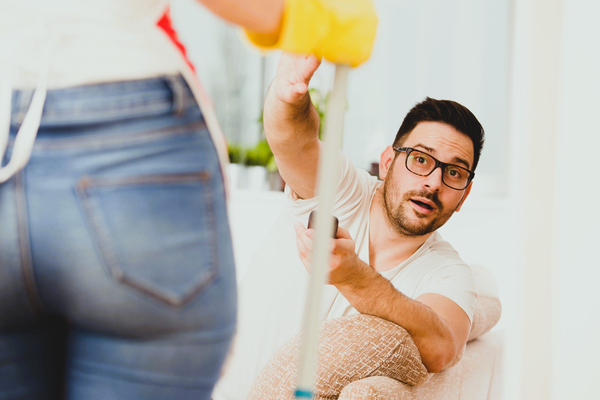 A man sitting on a sofa looks surprised while reaching for a mop held by someone standing. The person standing wears jeans and yellow cleaning gloves. The setting appears to be a living room with light-colored furniture.