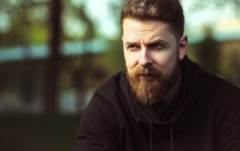 A bearded man in a black hoodie looks thoughtfully into the distance. The background is blurred with hints of greenery and a bright sky, creating a serene outdoor setting.