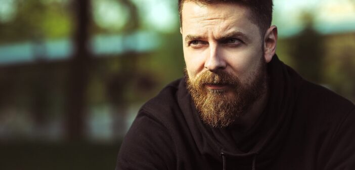 A bearded man in a black hoodie looks thoughtfully into the distance. The background is blurred with hints of greenery and a bright sky, creating a serene outdoor setting.