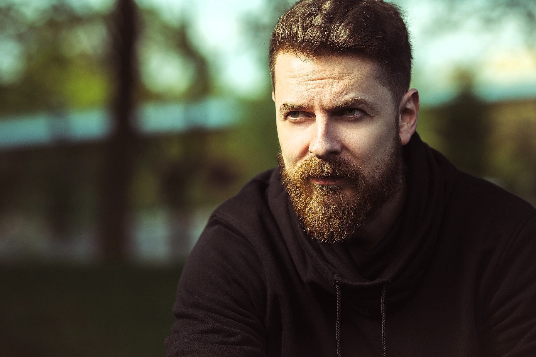 A bearded man in a black hoodie looks thoughtfully into the distance. The background is blurred with hints of greenery and a bright sky, creating a serene outdoor setting.