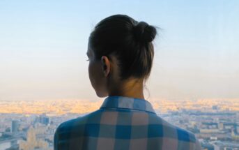 A person with their hair in a bun, wearing a blue and white checkered shirt, stands indoors facing a large window. They overlook a vast cityscape bathed in warm sunlight under a clear blue sky.