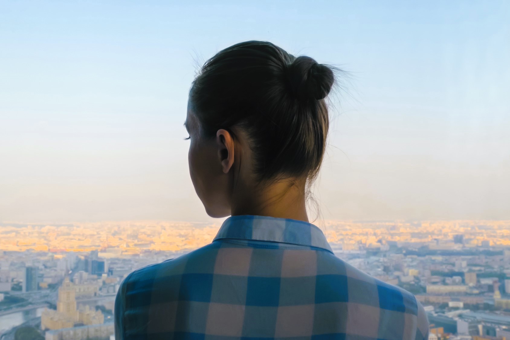 A person with their hair in a bun, wearing a blue and white checkered shirt, stands indoors facing a large window. They overlook a vast cityscape bathed in warm sunlight under a clear blue sky.