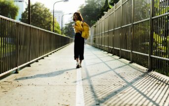 A woman wearing a yellow jacket and backpack walks on a pedestrian bridge surrounded by metal railings. The sun casts shadows of the railings on the walkway. Trees and buildings are visible in the background.