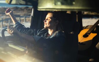 A woman sits in a car with her eyes closed, smiling and enjoying the sunlight streaming in. Her left hand is raised, and there's an acoustic guitar on the seat behind her. The scene suggests a relaxed, sunny day.