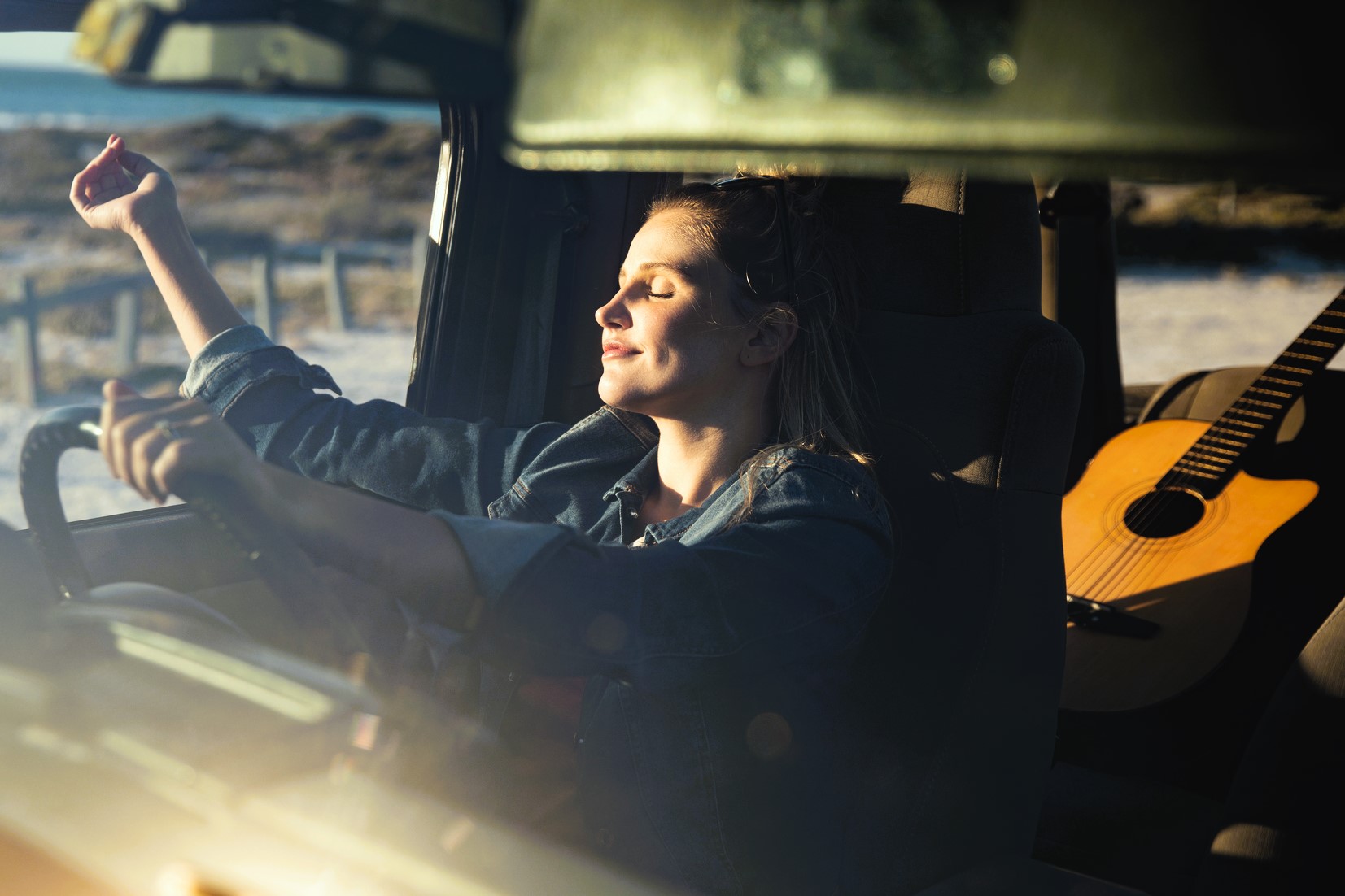 A woman sits in a car with her eyes closed, smiling and enjoying the sunlight streaming in. Her left hand is raised, and there's an acoustic guitar on the seat behind her. The scene suggests a relaxed, sunny day.