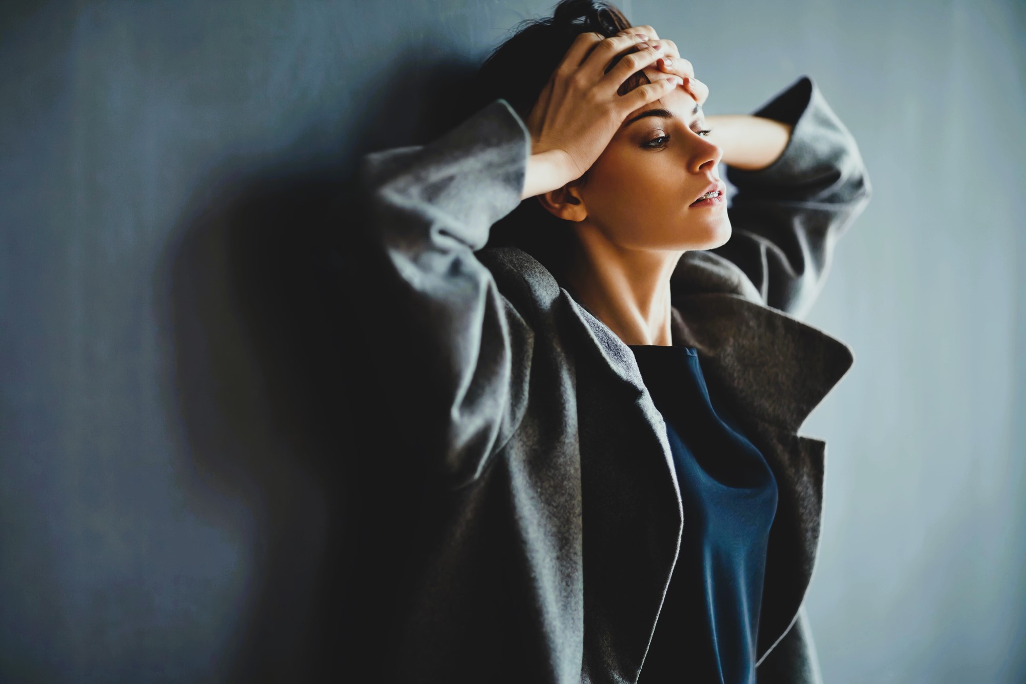 A person stands against a dark background, wearing a gray coat and a dark shirt. They have short, dark hair and are holding their head with both hands, gazing upward thoughtfully. The lighting creates a soft, moody atmosphere.