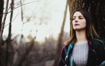 Woman with long hair wearing a sweater and plaid scarf leans against a tree in a wooded area. The background is blurred, with bare branches and soft sunlight filtering through, creating a serene, contemplative atmosphere.