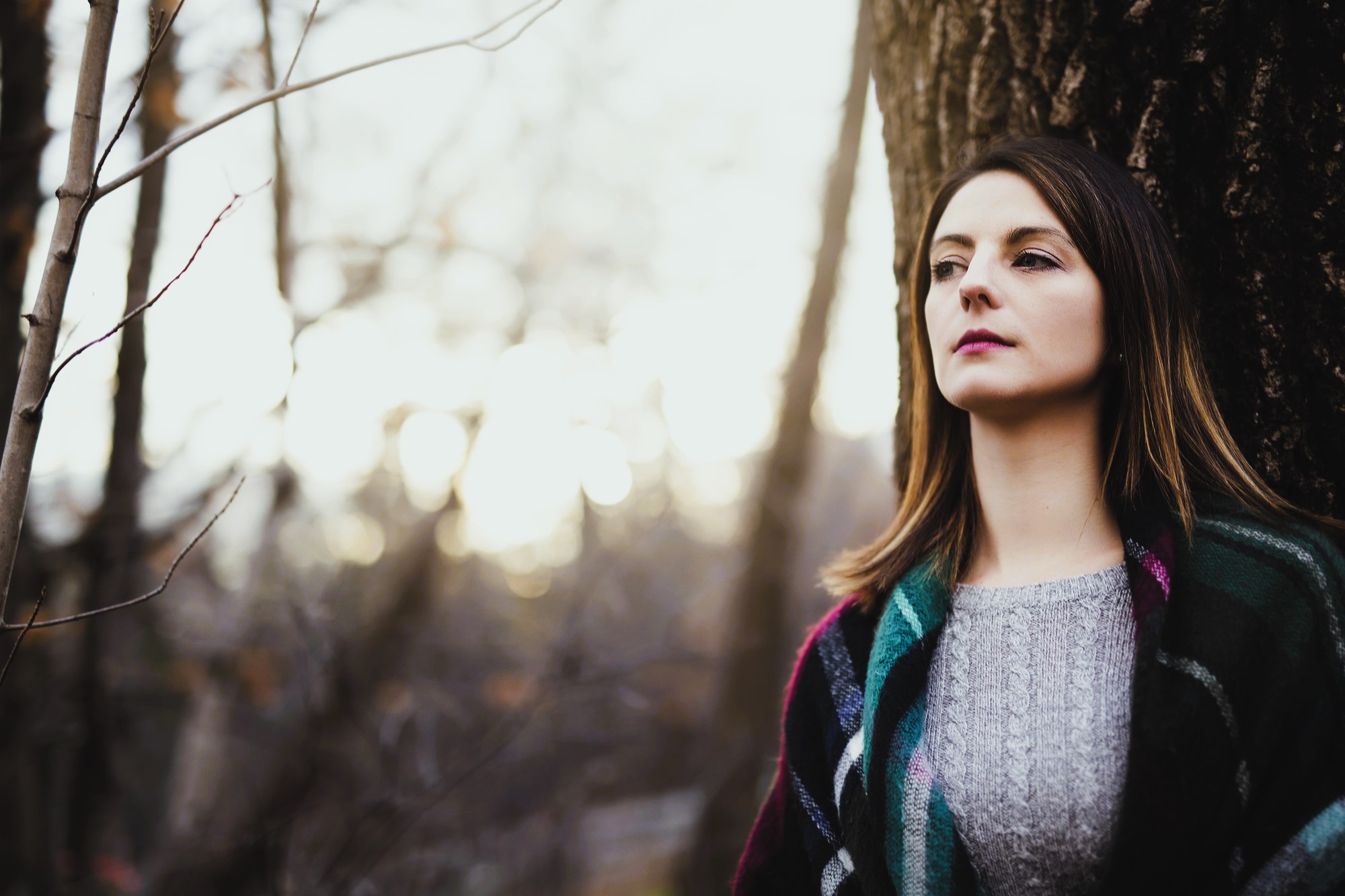 Woman with long hair wearing a sweater and plaid scarf leans against a tree in a wooded area. The background is blurred, with bare branches and soft sunlight filtering through, creating a serene, contemplative atmosphere.