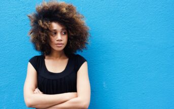 Person with curly hair standing against a bright blue wall, arms crossed and looking to the side. Wearing a black top.