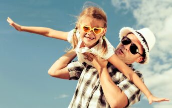 Man wearing a straw hat playfully lifts a smiling girl with heart-shaped sunglasses, both wearing casual summer clothing. They pose against a bright blue sky with a few clouds, creating a joyful, sun-filled scene.