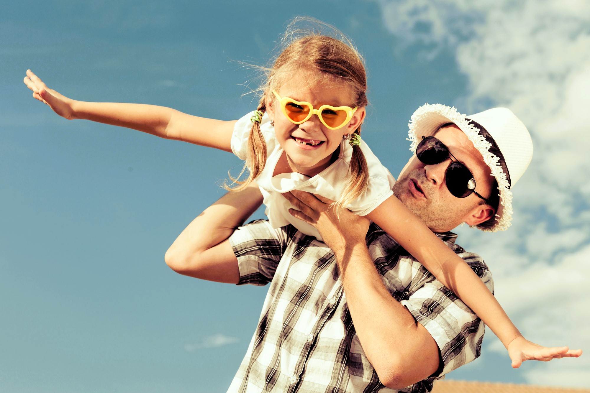 Man wearing a straw hat playfully lifts a smiling girl with heart-shaped sunglasses, both wearing casual summer clothing. They pose against a bright blue sky with a few clouds, creating a joyful, sun-filled scene.