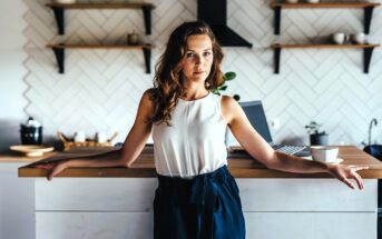 A woman with wavy brown hair stands in a modern kitchen, leaning against a wooden countertop. She wears a white sleeveless top and dark pants. There are shelves and kitchen items in the background, with a cup on the counter beside her.