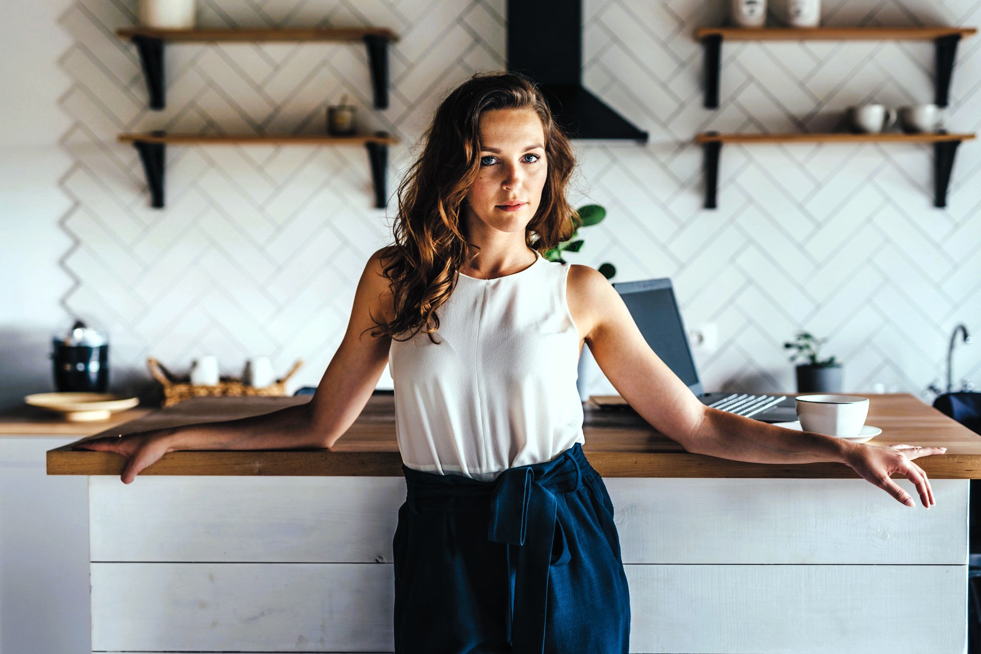 A woman with wavy brown hair stands in a modern kitchen, leaning against a wooden countertop. She wears a white sleeveless top and dark pants. There are shelves and kitchen items in the background, with a cup on the counter beside her.