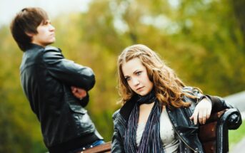 A woman with long curly hair sits on a bench, looking thoughtful, while a man stands in the background with arms crossed. Both are wearing black leather jackets. The scene is set outdoors with blurred greenery in the background.