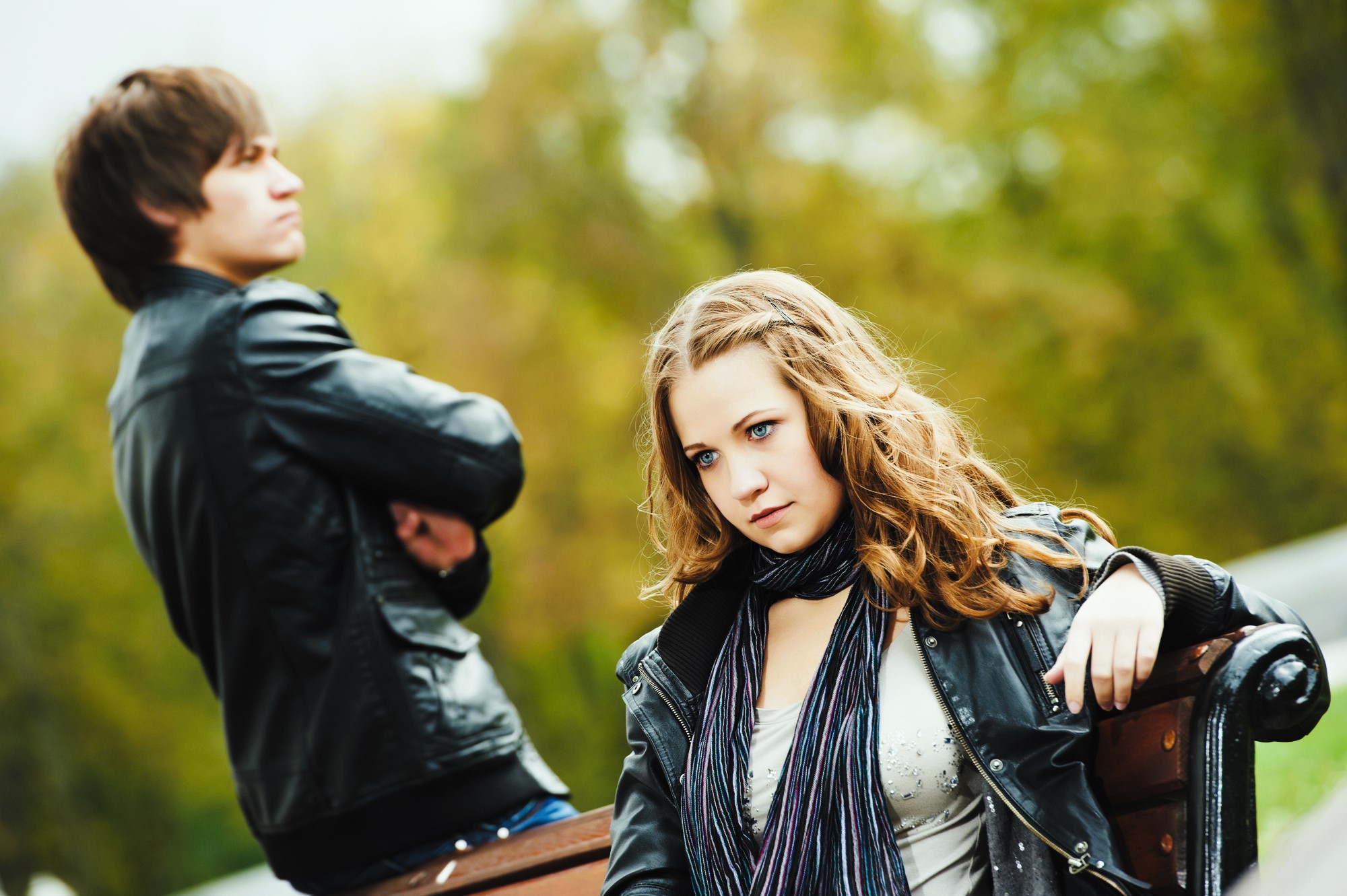 A woman with long curly hair sits on a bench, looking thoughtful, while a man stands in the background with arms crossed. Both are wearing black leather jackets. The scene is set outdoors with blurred greenery in the background.