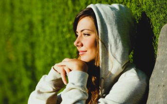 Woman in a gray hoodie leans against a wall, gazing thoughtfully to the side with a serene expression. Sunlight highlights her features, and the background is a lush green hedge.