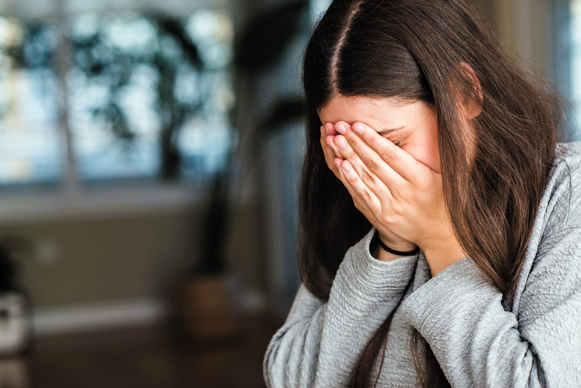 A person with long brown hair is sitting indoors, covering their face with both hands. They are wearing a gray top. The background is softly blurred, showing a window with light coming through and some indoor plants.