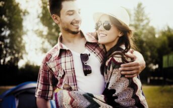 A smiling couple enjoying a sunny day outdoors. The man is wearing a plaid shirt and sunglasses, with an arm around the woman. The woman is wearing a hat, sunglasses, and a colorful cardigan. They appear happy and relaxed, with trees in the background.