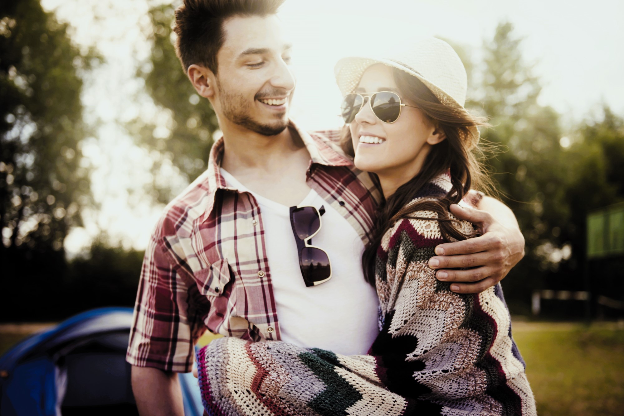 A smiling couple enjoying a sunny day outdoors. The man is wearing a plaid shirt and sunglasses, with an arm around the woman. The woman is wearing a hat, sunglasses, and a colorful cardigan. They appear happy and relaxed, with trees in the background.