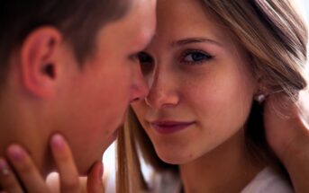 A close-up of two people facing each other intimately. The person on the right has long hair, and is looking at the camera with a gentle expression. The person on the left is blurry, focusing on the person on the right.