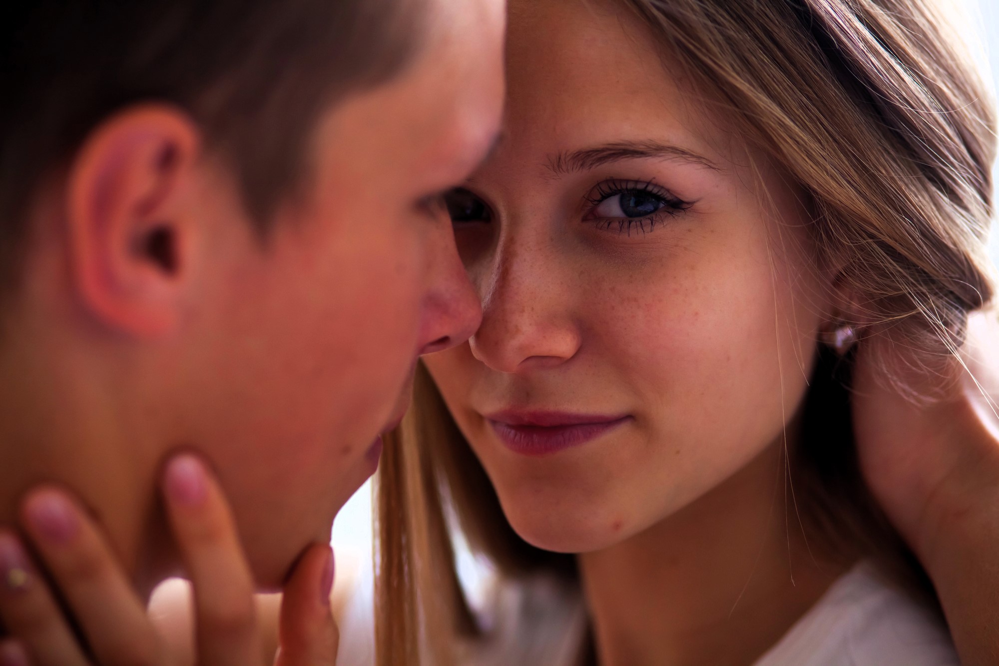 A close-up of two people facing each other intimately. The person on the right has long hair, and is looking at the camera with a gentle expression. The person on the left is blurry, focusing on the person on the right.