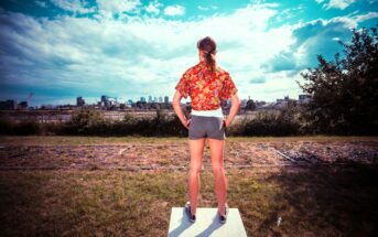 A person with long hair stands on a platform outdoors, facing a city skyline. They wear a red patterned shirt, gray shorts, and sneakers, hands on hips. The sky is partly cloudy, and the foreground includes patches of grass and shrubs.