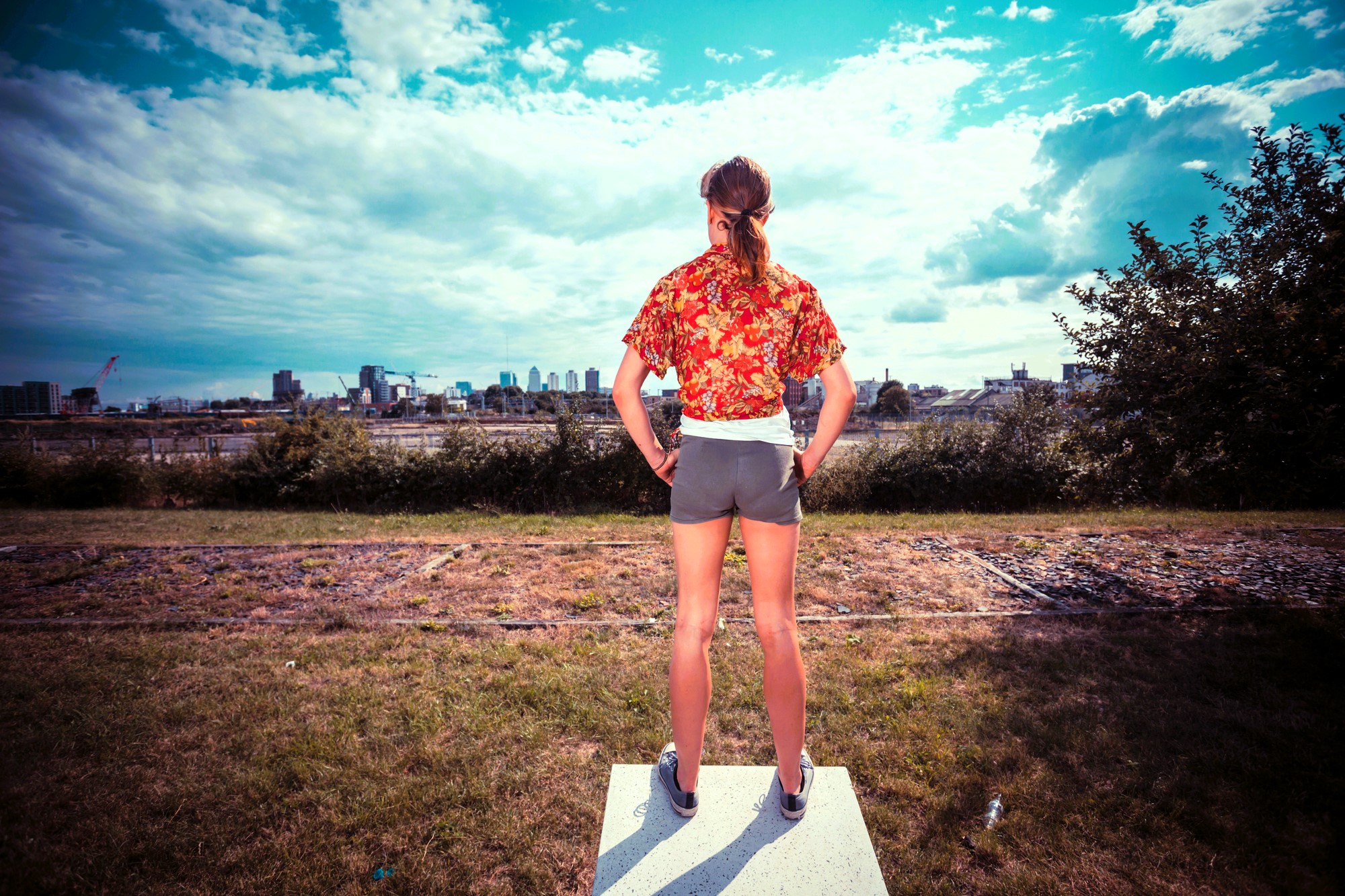 A person with long hair stands on a platform outdoors, facing a city skyline. They wear a red patterned shirt, gray shorts, and sneakers, hands on hips. The sky is partly cloudy, and the foreground includes patches of grass and shrubs.