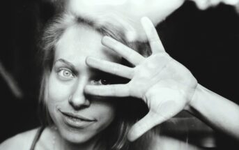 Black and white image of a person smiling while looking through a glass pane. They hold their hand up with fingers spread across their face, creating an interesting pattern of light and shadow.