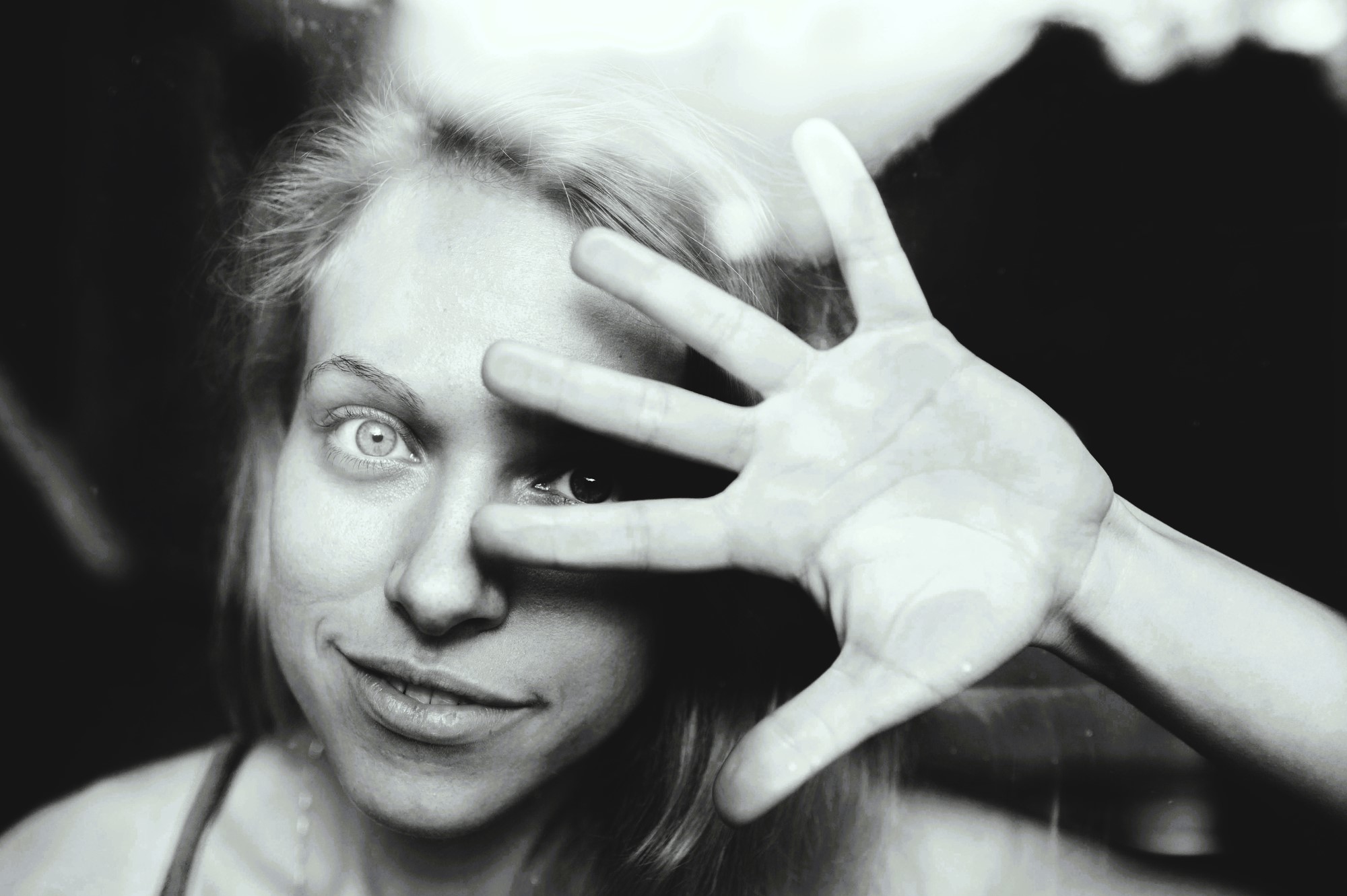 Black and white image of a person smiling while looking through a glass pane. They hold their hand up with fingers spread across their face, creating an interesting pattern of light and shadow.