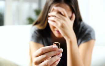 A woman with long hair sits on a couch, holding a ring in her right hand. Her left hand covers her face, indicating distress or contemplation. She wears a gray shirt, and the background is softly blurred.