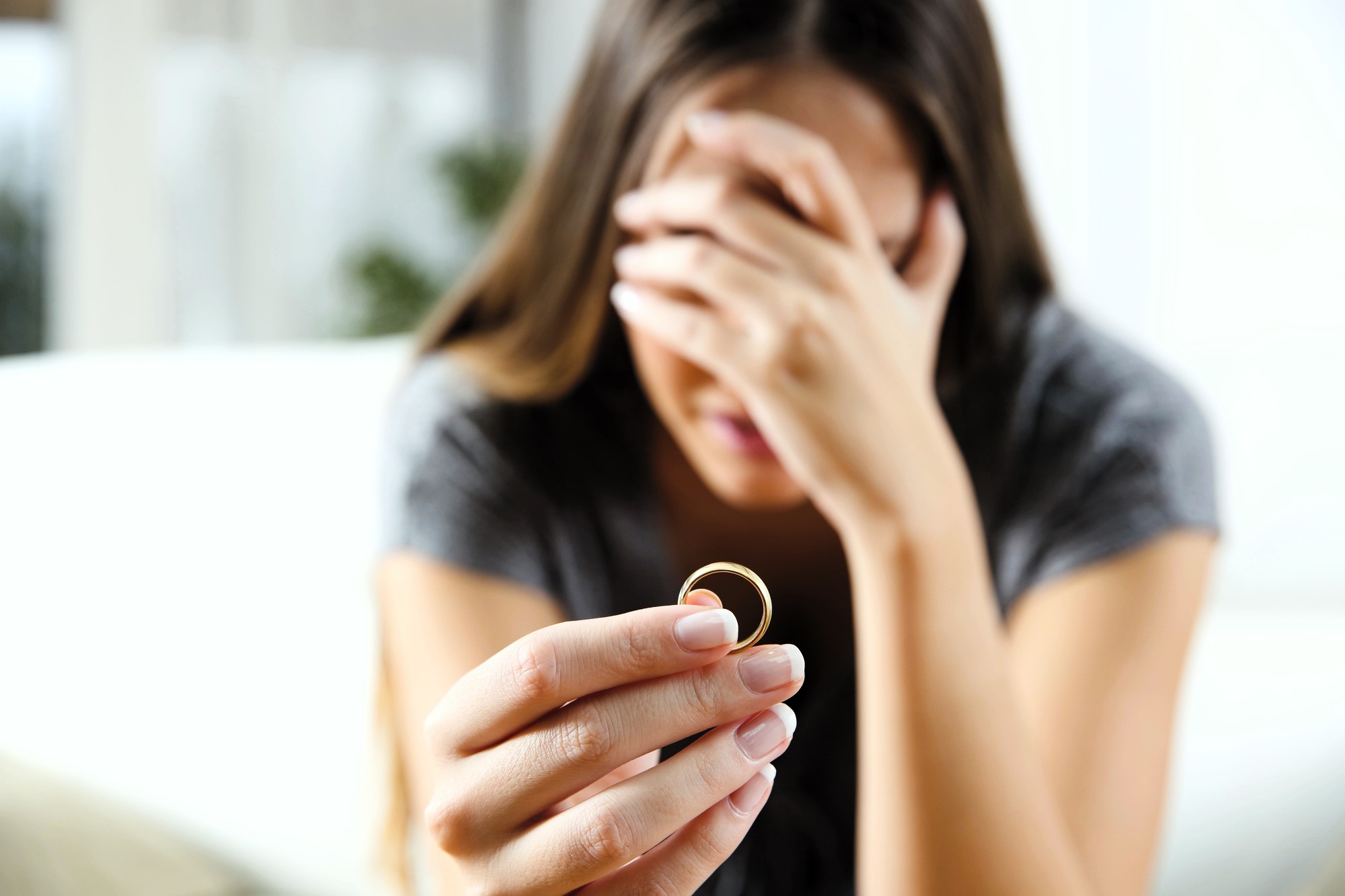 A woman with long hair sits on a couch, holding a ring in her right hand. Her left hand covers her face, indicating distress or contemplation. She wears a gray shirt, and the background is softly blurred.