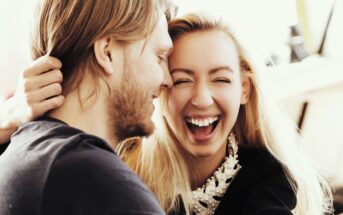 A man and a woman are joyfully laughing together, facing each other closely. The woman has her eyes closed with a wide smile, wearing a pearl-adorned garment. The man, with long hair and beard, is smiling. The background is softly blurred.