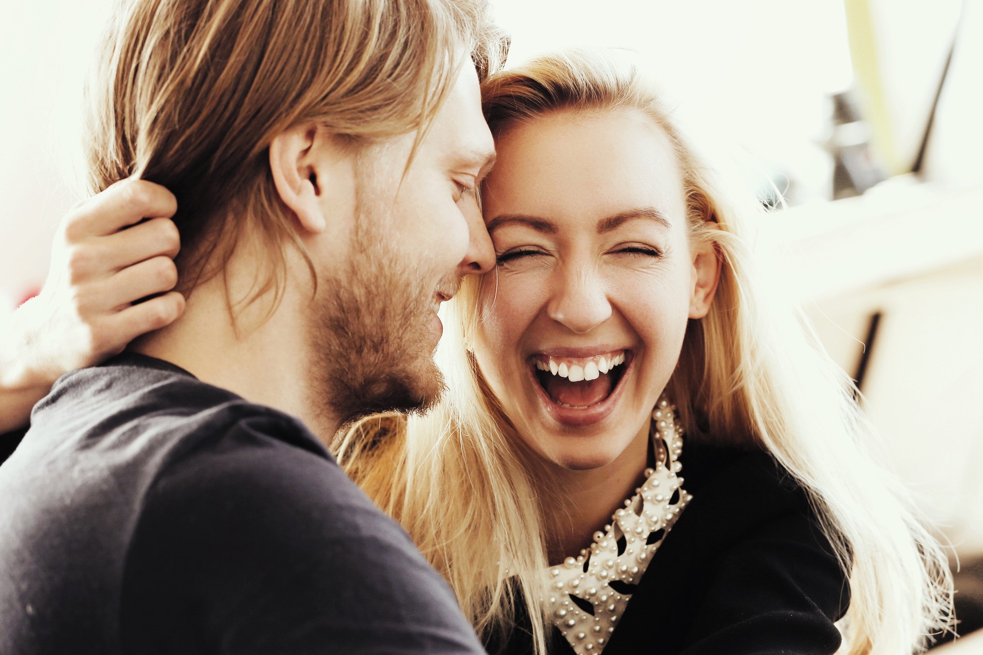 A man and a woman are joyfully laughing together, facing each other closely. The woman has her eyes closed with a wide smile, wearing a pearl-adorned garment. The man, with long hair and beard, is smiling. The background is softly blurred.