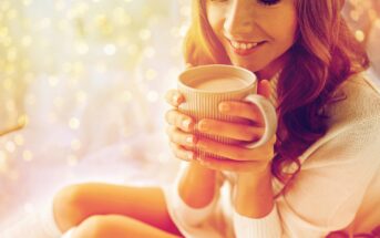 Woman smiling, holding a ribbed mug close to her face. She's wearing a cozy sweater, surrounded by a warm, softly blurred golden bokeh background, creating a serene and comforting atmosphere.