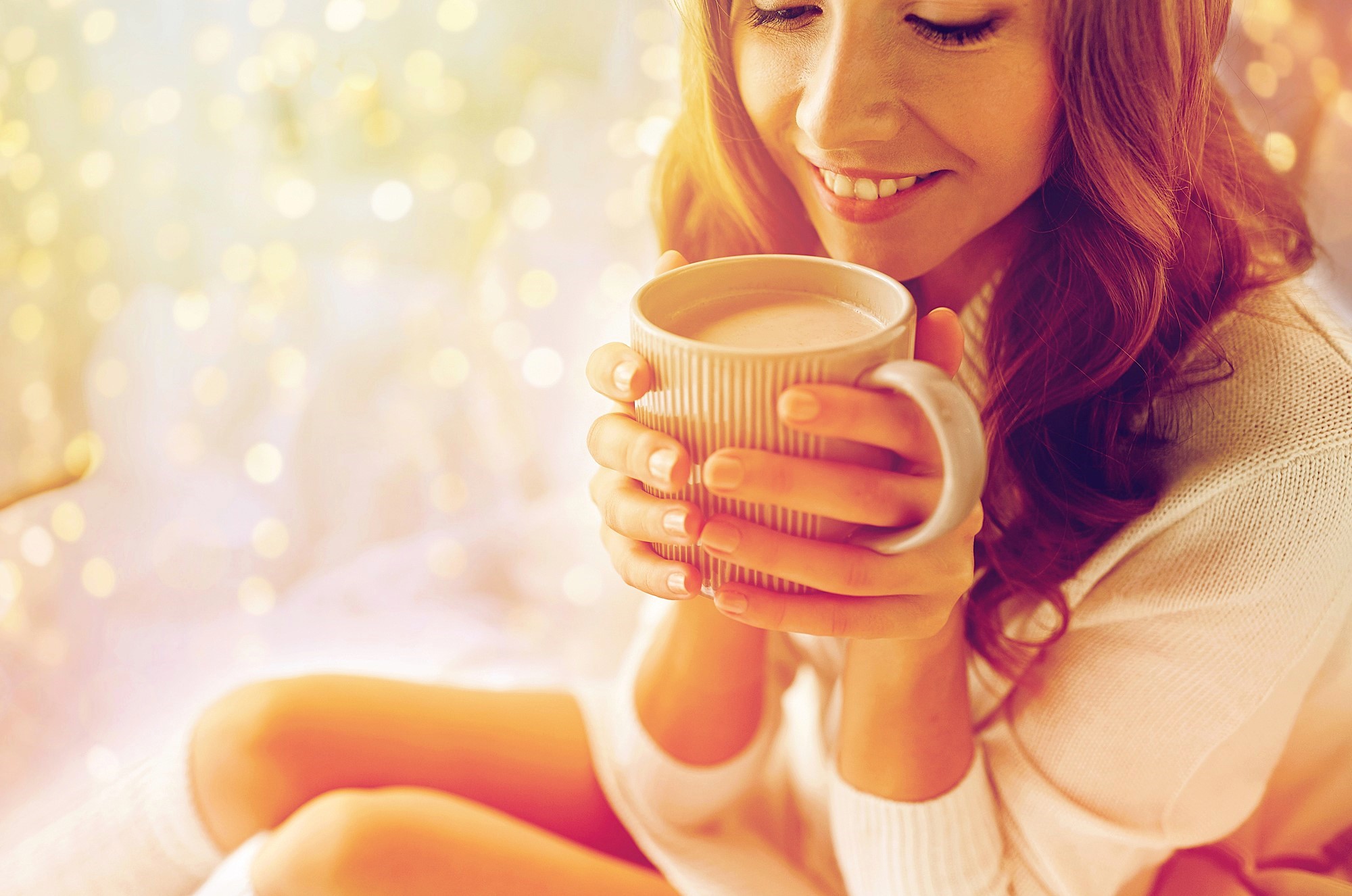 Woman smiling, holding a ribbed mug close to her face. She's wearing a cozy sweater, surrounded by a warm, softly blurred golden bokeh background, creating a serene and comforting atmosphere.