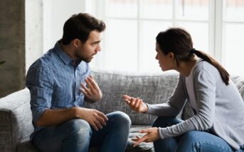 A man and a woman sit on a gray sofa, engaged in a heated conversation. The man gestures with his left hand, wearing a blue checkered shirt and jeans. The woman faces him, gesturing with her right hand, wearing a gray sweater and jeans.