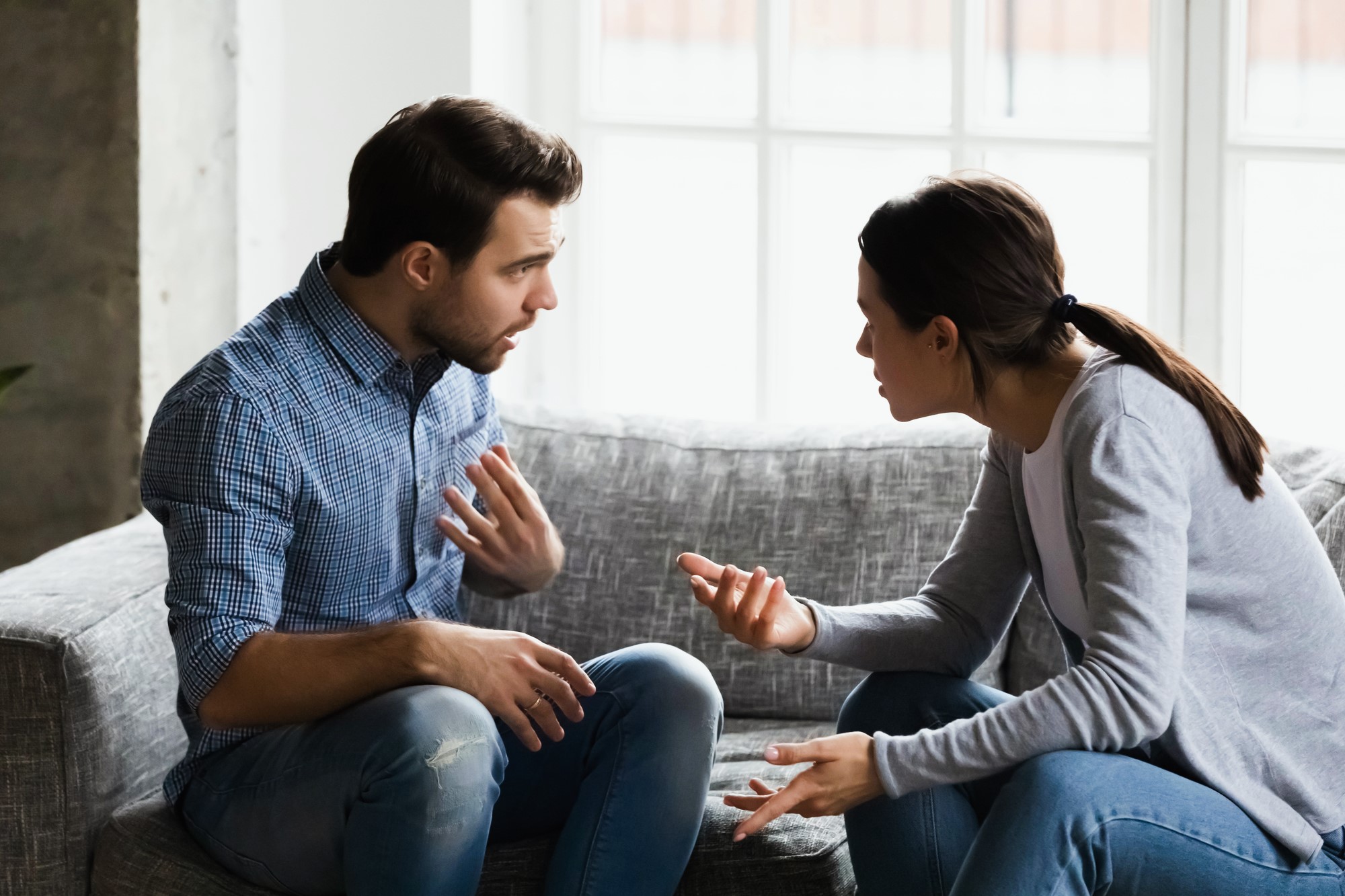 A man and a woman sit on a gray sofa, engaged in a heated conversation. The man gestures with his left hand, wearing a blue checkered shirt and jeans. The woman faces him, gesturing with her right hand, wearing a gray sweater and jeans.