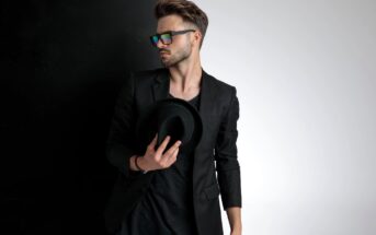 A man with styled hair wears a dark blazer, T-shirt, and rainbow-reflective sunglasses. He holds a black hat in one hand. The background transitions from black on the left to white on the right.