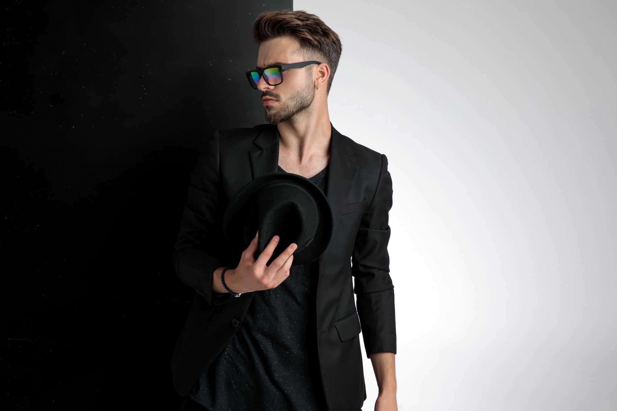 A man with styled hair wears a dark blazer, T-shirt, and rainbow-reflective sunglasses. He holds a black hat in one hand. The background transitions from black on the left to white on the right.