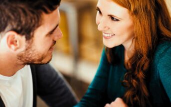 A man and woman sit closely, smiling and looking into each other's eyes. The woman has long red hair and wears a teal sweater, while the man has short dark hair and wears a grey cardigan over a white shirt. The background is softly blurred.