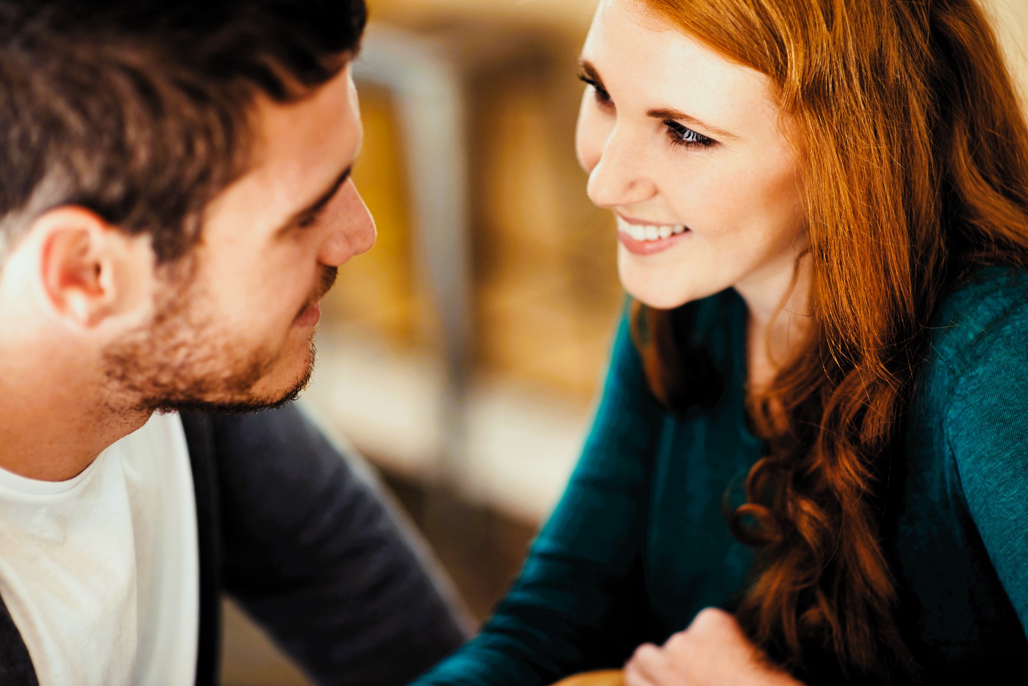 A man and woman sit closely, smiling and looking into each other's eyes. The woman has long red hair and wears a teal sweater, while the man has short dark hair and wears a grey cardigan over a white shirt. The background is softly blurred.