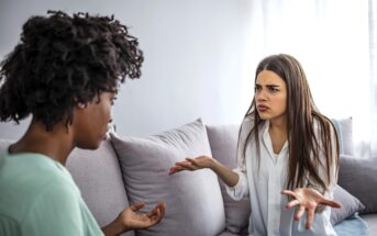 Two women are sitting on a couch in a living room. The woman on the right has long hair and appears frustrated, gesturing with her hands. The woman on the left has curly hair and is listening attentively. They appear to be having a serious conversation.