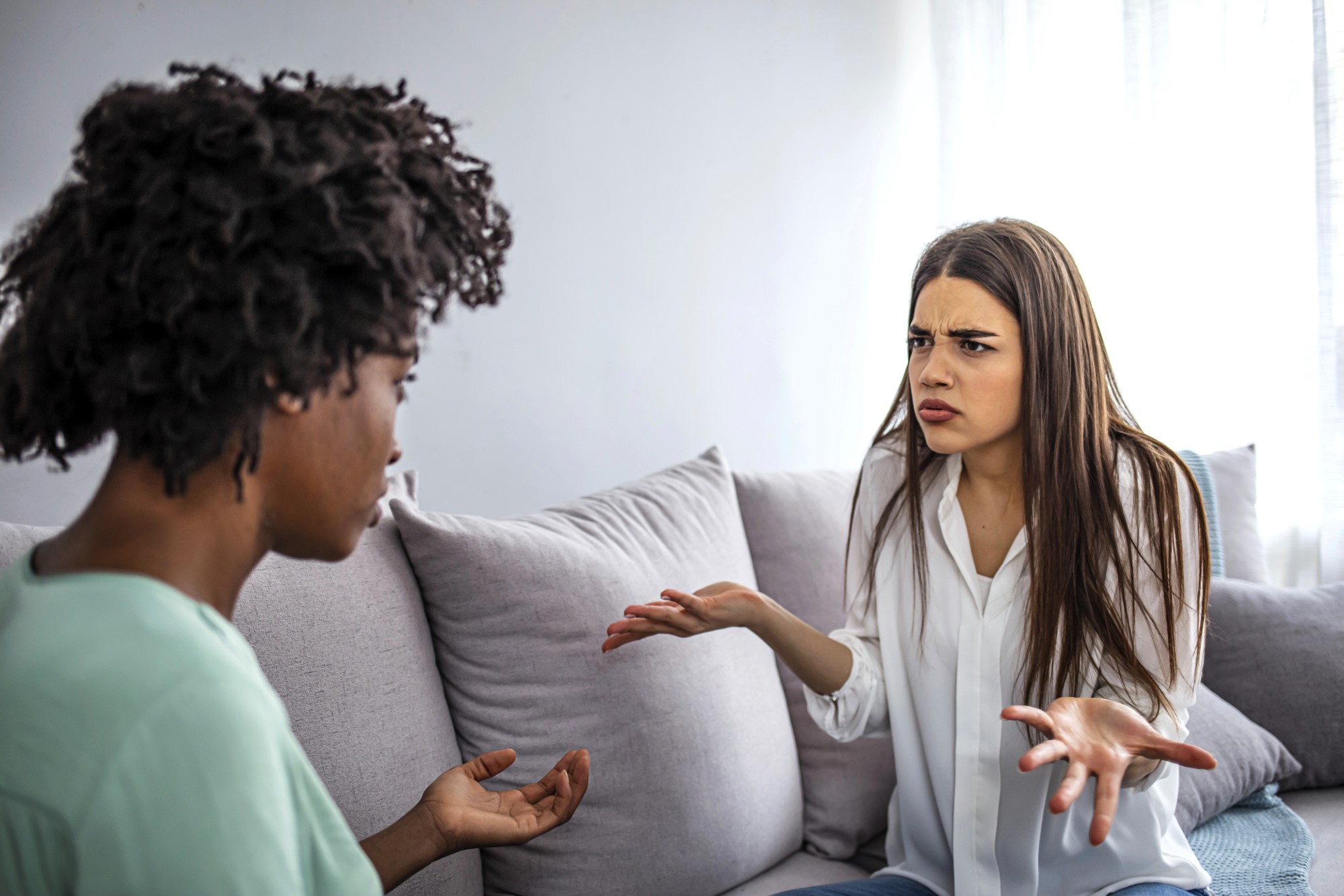 Two women are sitting on a couch in a living room. The woman on the right has long hair and appears frustrated, gesturing with her hands. The woman on the left has curly hair and is listening attentively. They appear to be having a serious conversation.